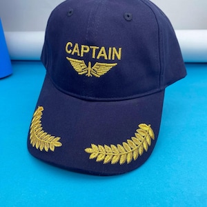 May include: A navy blue baseball cap with the word "CAPTAIN" embroidered in gold, along with a gold emblem of wings and a plane. Gold leaf embroidery adorns the brim.