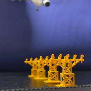 Op de afbeelding: Een miniatuur geel luchthavenverlichtingssysteem met een modelvliegtuig op de achtergrond. Het verlichtingssysteem heeft een reeks gele torens met rode lichten. Het vliegtuig is wit met zwarte motoren en is in vlucht.