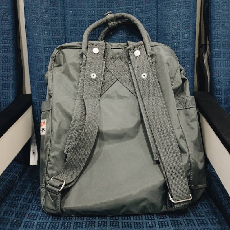 Puede incluir: Una mochila de lona gris con correas ajustables y un asa superior. La mochila tiene un peque&ntilde;o parche con el texto "SA 39" en la parte delantera.