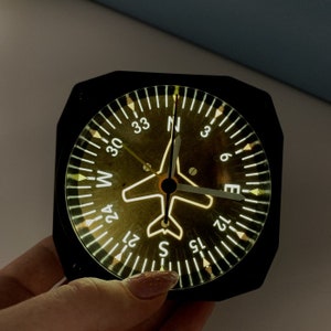 Puede incluir: Una brújula negra con una silueta de avión blanca en el centro. La cara de la brújula está marcada con números del 0 al 36 y las direcciones cardinales N, S, E y W. La brújula está iluminada con una luz amarilla cálida.