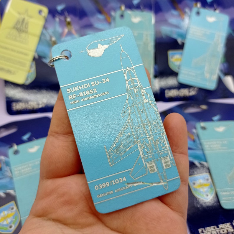 May include: A light blue metal keychain with a white outline of a Sukhoi SU-34 fighter jet. The keychain has the text "SUKHOI SU-34", "RF-81852", "MSN: 4160662810801", "0399/1034", and "GENUINE AIRCRAFT".