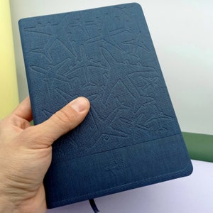 May include: A dark blue notebook with a textured cover featuring an embossed pattern of airplanes. The notebook has a simple, modern design.