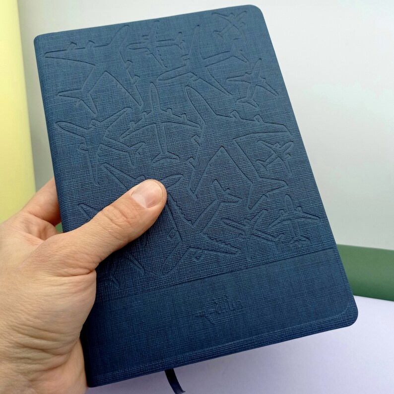 May include: A dark blue notebook with a textured cover featuring an embossed pattern of airplanes. The notebook has a simple, modern design.