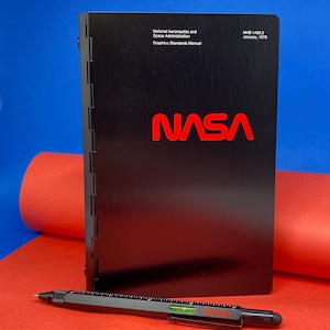 May include: A black, metal-bound book with the title "National Aeronautics and Space Administration Graphics Standards Manual" and the NASA logo in red. The book is on a red surface with a blue surface in the background. The text "NHB 1430.2 January, 1978" is printed on the top right corner of the book.
