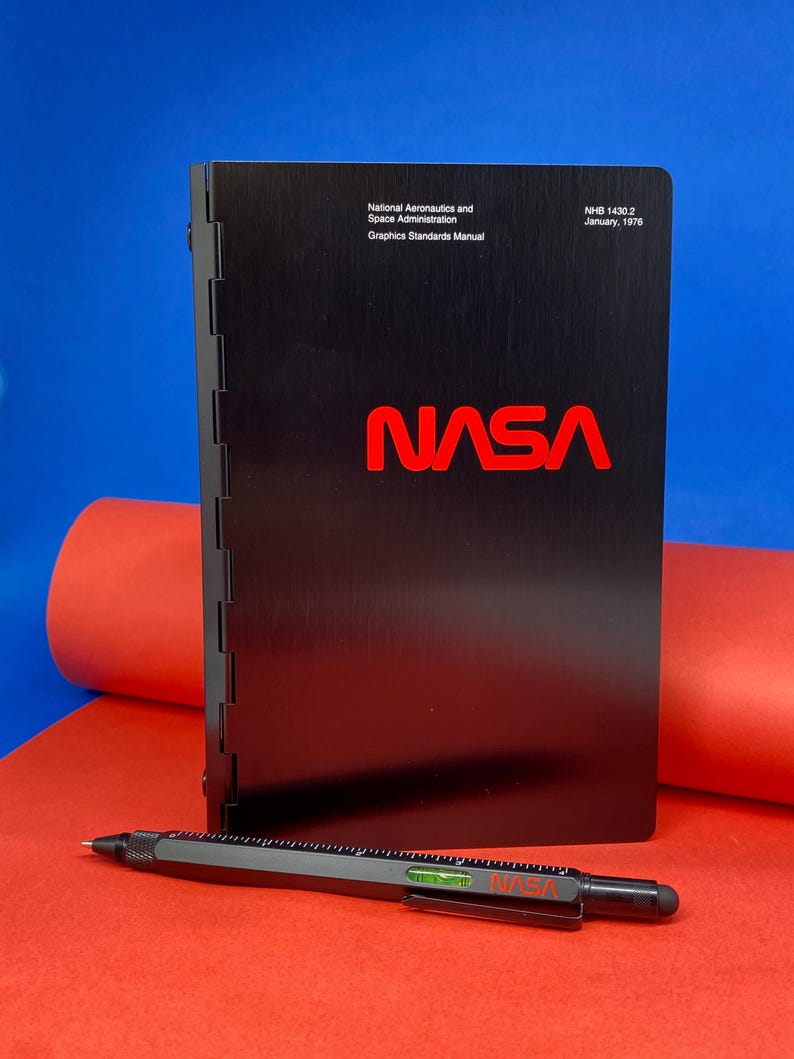 May include: A black, metal-bound book with the title "National Aeronautics and Space Administration Graphics Standards Manual" and the NASA logo in red. The book is on a red surface with a blue surface in the background. The text "NHB 1430.2 January, 1978" is printed on the top right corner of the book.