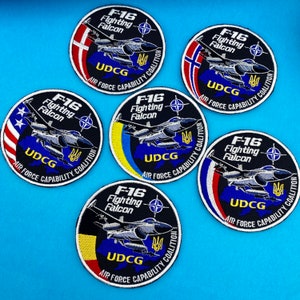 May include: Six round black and white embroidered patches with the text "F-16 Fighting Falcon" and "UDCG AIR FORCE CAPABILITY COALITION". Each patch features a different flag in the background, including the American, Ukrainian, Danish, and Norwegian flags.