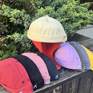 Puede incluir: Una colección de gorros tipo docker sin visera en varios colores, incluyendo crema, rojo, negro, rosa, azul oscuro, lavanda, verde oliva, gris y amarillo. Cada gorro presenta un pequeño icono de avión bordado en el borde y un botón superior.