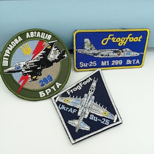 Può includere: Tre toppe ricamate con disegni di aerei militari. Una è circolare con testo ucraino e un jet, un'altra è rettangolare con sfondo blu e la parola "Frogfoot", e la terza è a forma di diamante con un jet e il testo "UkrAF Su-25".