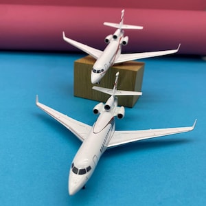 Pode incluir: Dois modelos de aviões brancos com detalhes em vermelho e preto são exibidos em uma superfície azul. Um avião está posicionado em um pequeno bloco de madeira, enquanto o outro está na frente. Os aviões têm asas e caudas detalhadas.