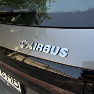 以下が含まれることがあります： 車のリア部分のクローズアップ。クロムメッキされた「AIRBUS」エンブレムが特徴です。エンブレムはナンバープレートエリアの上に配置されています。車のボディは明るいグレーで、リアウィンドウが見えます。