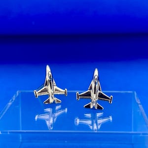 May include: A pair of silver-colored airplane-shaped cufflinks. The cufflinks are displayed on a clear, rectangular surface against a vibrant blue background. The airplanes have detailed wings and tails.