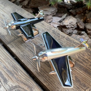 Op de afbeelding: Twee modelvliegtuigen met zilveren en zwarte carrosserieën, zilveren propellers en chromen accenten worden tentoongesteld op een houten oppervlak. De vleugels hebben een gestreept ontwerp. De vliegtuigen zijn miniatuurreplica's van vintage vliegtuigen.