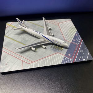 Pode incluir: Um modelo de avião branco e azul com o logotipo EL AL está em um diorama de pista cinza e vermelho. O modelo tem quatro motores e asas detalhadas. O diorama tem marcações de pista e taxiways pintadas.