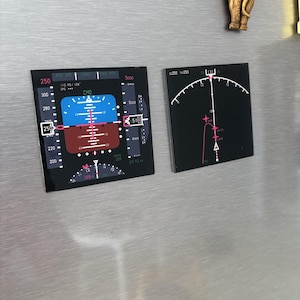 Puede incluir: Dos imanes cuadrados negros con diseños de paneles de instrumentos de aviación. Uno presenta una pantalla azul y roja con números y texto, el otro un indicador curvo con marcas rosas. También se ve una pequeña figura de elefante.