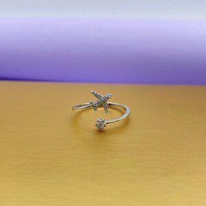 Puede incluir: Un anillo plateado con forma de avi&oacute;n con un dise&ntilde;o envolvente. El avi&oacute;n y el peque&ntilde;o adorno floral est&aacute;n incrustados con peque&ntilde;as piedras preciosas transparentes. El anillo est&aacute; sobre una superficie dorada con fondo morado.