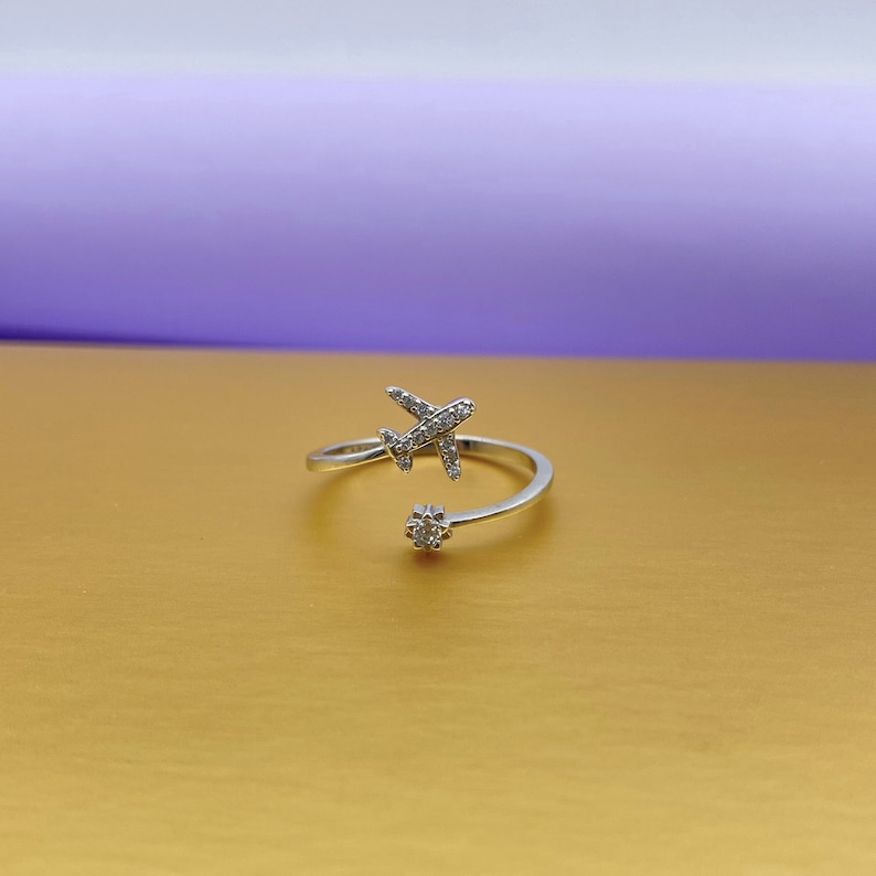 Puede incluir: Un anillo plateado con forma de avi&oacute;n con un dise&ntilde;o envolvente. El avi&oacute;n y el peque&ntilde;o adorno floral est&aacute;n incrustados con peque&ntilde;as piedras preciosas transparentes. El anillo est&aacute; sobre una superficie dorada con fondo morado.