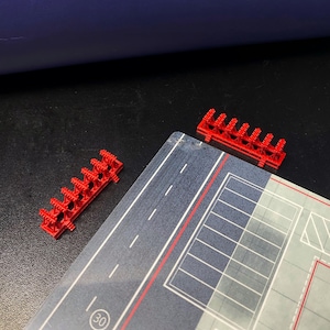 May include: Red plastic toy barricades are placed near a printed play mat with a road and parking lot design. The mat has a speed limit sign of 30. The toy barricades are small and have a series of vertical bars.