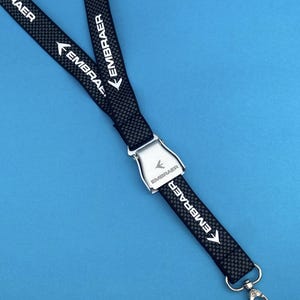 Könnte beinhalten: Schwarzes Schlüsselband mit dem weißen Schriftzug "EMBRAER" und einem Pfeil-Grafik. Das Schlüsselband hat ein Karbonfasermuster und einen silberfarbenen Metallverschluss. Der Hintergrund ist einfarbig blau.