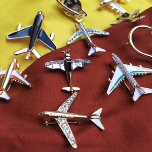 May include: Various airplane brooches in gold, silver, and black, some with rhinestone detailing and white accents. One brooch features the text "Good Luck". The brooches are displayed against a red and yellow backdrop.