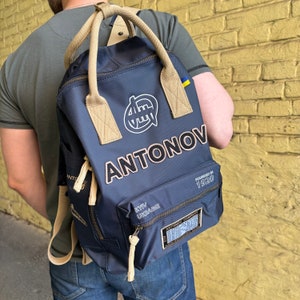 May include: Navy blue backpack with the name "ANTONOV" in white and black lettering. The backpack has tan straps and handles, and a white logo. The text "KYIV UKRAINE" and "FOUNDED IN 1920" are also visible.