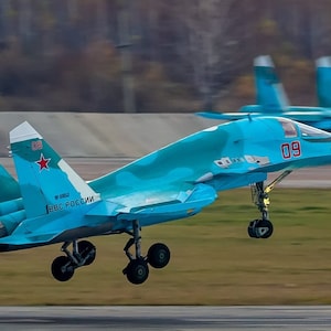 May include: A blue and white fighter jet with the number 09 on the side, taking off from a runway. The jet has a red star on the tail and the words "ВКС РОССИИ" on the side.