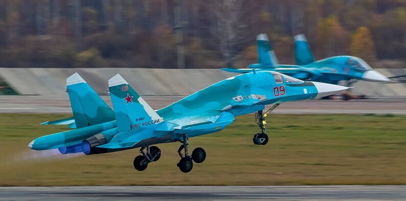 May include: A blue and white fighter jet with the number 09 on the side, taking off from a runway. The jet has a red star on the tail and the words "ВКС РОССИИ" on the side.