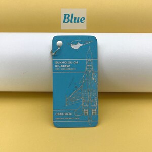 May include: A blue metal keychain with a silver ring. The keychain features a detailed illustration of a Sukhoi SU-34 fighter jet with the text "SUKHOI SU-34", "RF-81852", "MSN: 4160662810801", "0288/1034", and "GENUINE AIRCRAFT SKIN".