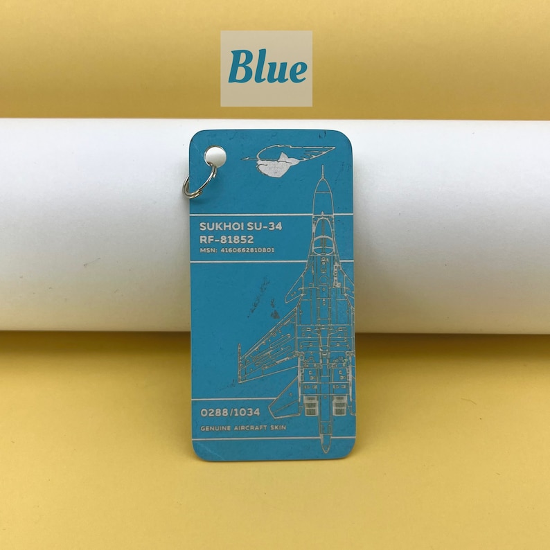 May include: A blue metal keychain with a silver ring. The keychain features a detailed illustration of a Sukhoi SU-34 fighter jet with the text "SUKHOI SU-34", "RF-81852", "MSN: 4160662810801", "0288/1034", and "GENUINE AIRCRAFT SKIN".