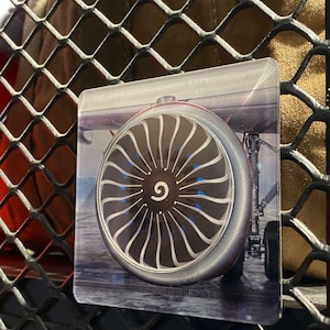 May include: A clear, rectangular acrylic magnet featuring a detailed image of an airplane engine turbine. The turbine has silver blades and a central black hub with a white '9' symbol. The background shows the plane's undercarriage.