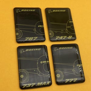 May include: Four black rectangular coasters with gold airplane designs. Each coaster features a different Boeing aircraft model: 787, 747-8, 737 MAX, and 777. The Boeing logo is at the top of each coaster.