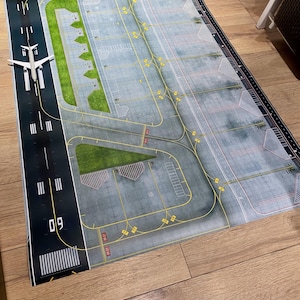 May include: A detailed airport play mat with a runway, taxiways, and parking areas. A white toy airplane sits on the runway. The mat features green grass areas and yellow markings, creating a realistic airport scene.