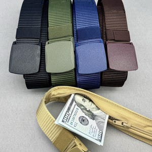 Può includere: Cinque cinture in nylon con fibbie in plastica nei colori nero, verde oliva, blu, marrone e beige. La cintura beige presenta uno scomparto nascosto con cerniera sul lato interno, contenente una banconota da cento dollari con testo visibile.