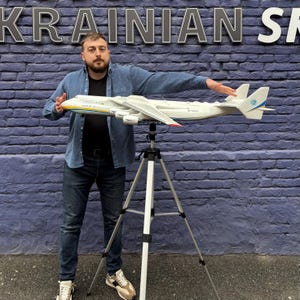 May include: A man is holding a white model airplane with blue and yellow accents. The airplane is on a tripod stand. The man is standing in front of a brick wall with the word "UKRAINIAN" written on it.