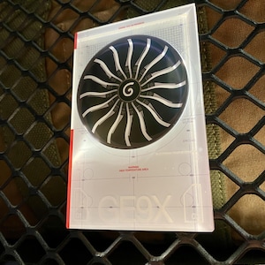 May include: A black and silver circular design with a 'G' in the center, resembling a turbine, is displayed on a clear plastic card. The card has the word 'GEOX' and the text 'WARNING HIGH TEMPERATURE AREA'.