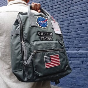 Puede incluir: Una mochila gris con un parche de la NASA, un parche de la bandera estadounidense y el texto "UNITED STATES" y "CASE TRAINING ROCKET SCIENCE TYPE R$2 SPEC No.42-20074-P".