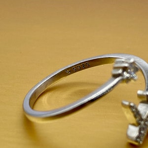 Puede incluir: Un anillo de plata con un dise&ntilde;o floral. El anillo tiene un dise&ntilde;o calado delicado con peque&ntilde;as piedras preciosas claras. La banda est&aacute; marcada con "S925". El anillo est&aacute; sobre un fondo dorado.