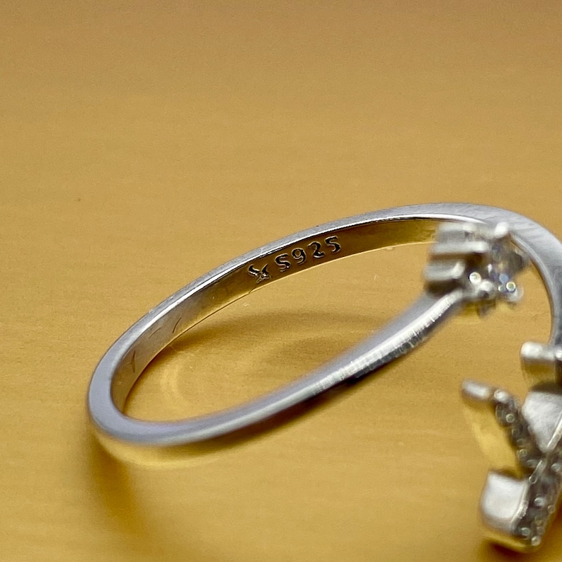 Puede incluir: Un anillo de plata con un dise&ntilde;o floral. El anillo tiene un dise&ntilde;o calado delicado con peque&ntilde;as piedras preciosas claras. La banda est&aacute; marcada con "S925". El anillo est&aacute; sobre un fondo dorado.