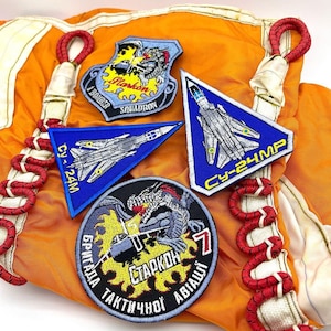 May include: Four embroidered patches on an orange fabric background. The patches are all different shapes and sizes. One patch is a blue and white fighter jet with the text "CY-24MP". Another patch is a yellow and black dragon with the text "Бригада Тактической Авиации" and "Старкон". The third patch is a blue and white fighter jet with the text "CY-24M". The fourth patch is a yellow and black fighter jet with the text "1 Border Squadron".