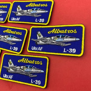 Op de afbeelding: Rechthoekige geborduurde patches met een blauwe achtergrond en een gele rand. Elke patch toont een grijs en blauw L-39 Albatros straalvliegtuig, de gele tekst Albatros en de witte tekst UkrAF en L-39 op een rode ondergrond.