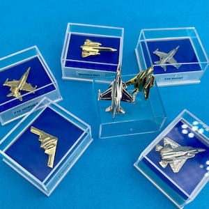 May include: A selection of miniature airplane pins displayed in clear cases. The pins are gold and silver, representing different aircraft models, such as the F18 Hornet and Blackbird SR71. Each case has a blue background.