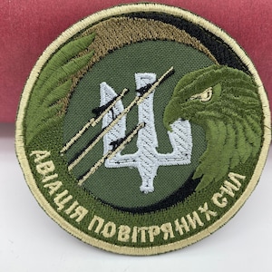 Może przedstawiać: Zielona naszywka haftowana z białym trójzębem i zielonym orłem. Tekst "АВІАЦІЯ ПОВІТРЯНИХ СИЛ" jest wyszyty wokół krawędzi naszywki.