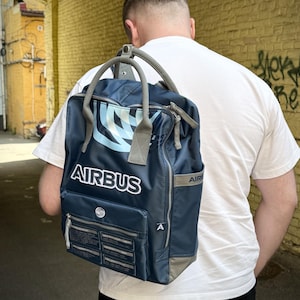 Puede incluir: Una mochila azul marino con el texto blanco AIRBUS en la parte frontal, un patrón gráfico en azul claro y blanco, asas superiores grises y un bolsillo delantero con varias etiquetas rectangulares que contienen texto blanco pequeño y un logo A.