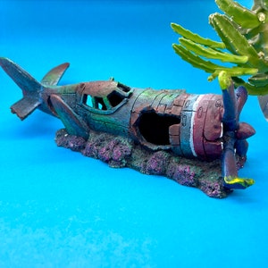 May include: A green, brown, and red model airplane wreck with a propeller, sitting on a bed of green and purple coral. The model is on a blue background.
