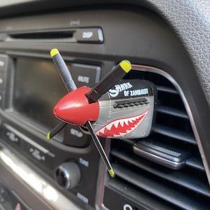 Puede incluir: Un ambientador de coche con forma de morro de avión de combate vintage, con un cono de hélice rojo, palas negras con puntas amarillas y un cuerpo plateado con un gráfico de boca de tiburón roja y blanca y el texto SHARK OF ZAMBALES impreso.