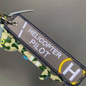 May include: A gray fabric keychain tag with the words "HELICOPTER PILOT" embroidered in white. The tag features a yellow and white helicopter landing pad symbol. A camouflage-patterned toy helicopter is visible.