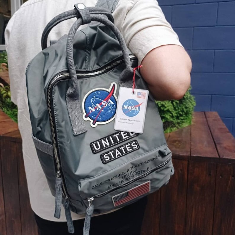 Puede incluir: Una mochila gris con un parche de la NASA y las palabras "United States" impresas en ella. La mochila tiene un bolsillo con cremallera en la parte delantera y un peque&ntilde;o parche de la bandera estadounidense en la parte inferior.