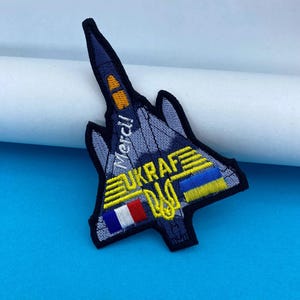 Pode incluir: Remendo bordado em forma de jato, com o texto "Merci!" e "UKRAF". O remendo inclui o brasão de armas ucraniano e as bandeiras da Ucrânia e da França. O remendo é principalmente azul marinho, amarelo e azul.