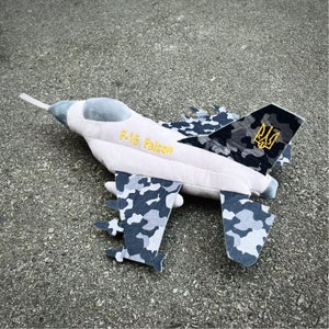 Puede incluir: Un avión de peluche gris y blanco con un patrón de camuflaje. El avión tiene las palabras "F-16 Falcon" impresas en el lateral y un símbolo de tridente ucraniano en la cola.