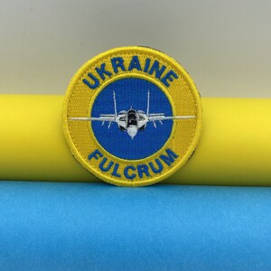 May include: A yellow and blue embroidered patch with a white fighter jet in the centre. The patch says "UKRAINE FULCRUM" around the jet.