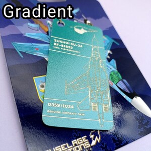May include: A teal blue keychain with a white outline of a Sukhoi SU-34 fighter jet. The keychain has the text "SUKHOI SU-34 RF-81852 MSN: 4160662810801 0359/1034 GENUINE AIRCRAFT SKIN" printed on it.